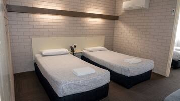 A hotel room with two beds, white brick walls, a ceiling-mounted air conditioner, and a wooden floor.