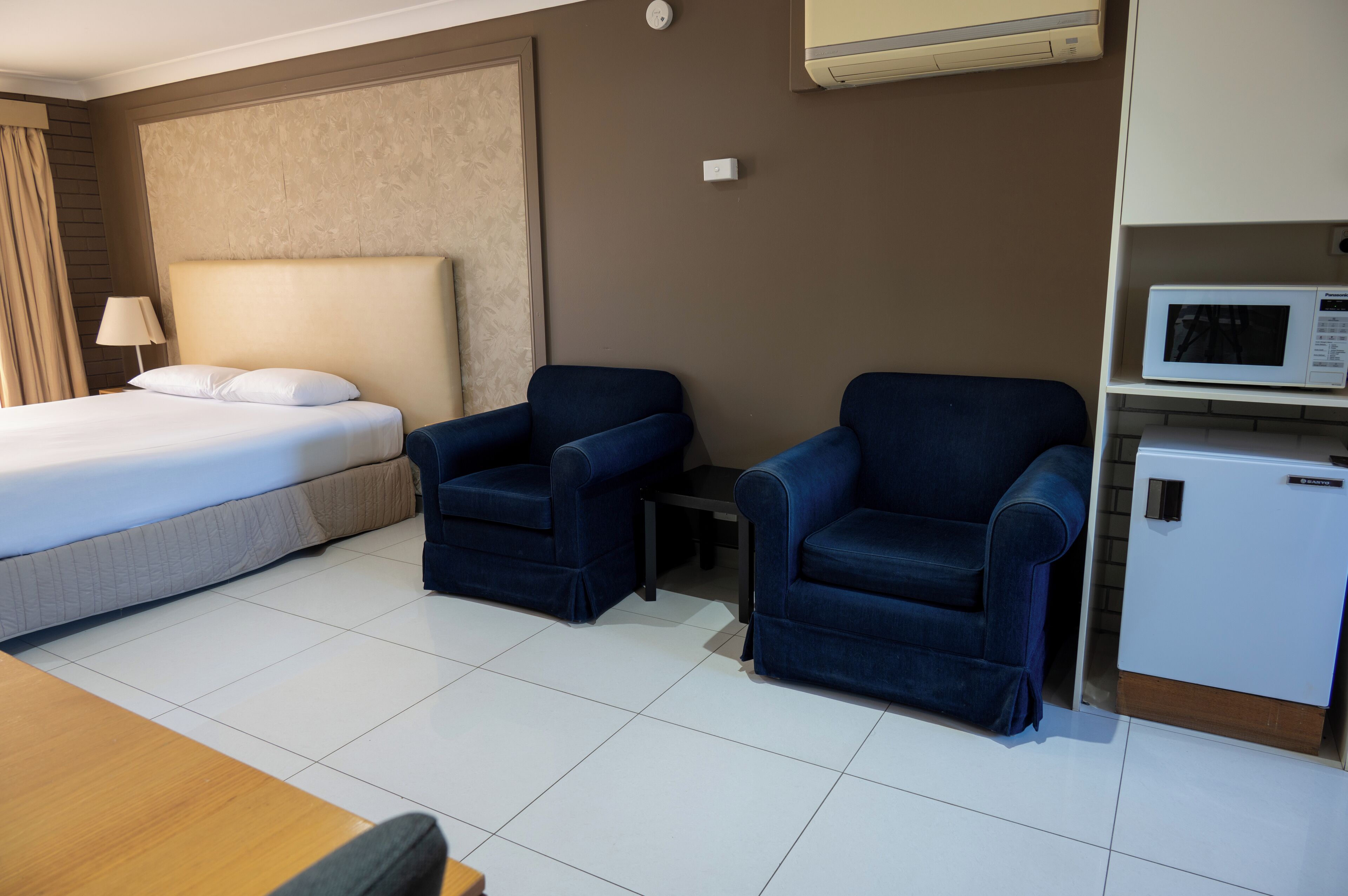 A hotel room with a bed, two blue armchairs, a small table, a microwave, and a refrigerator.