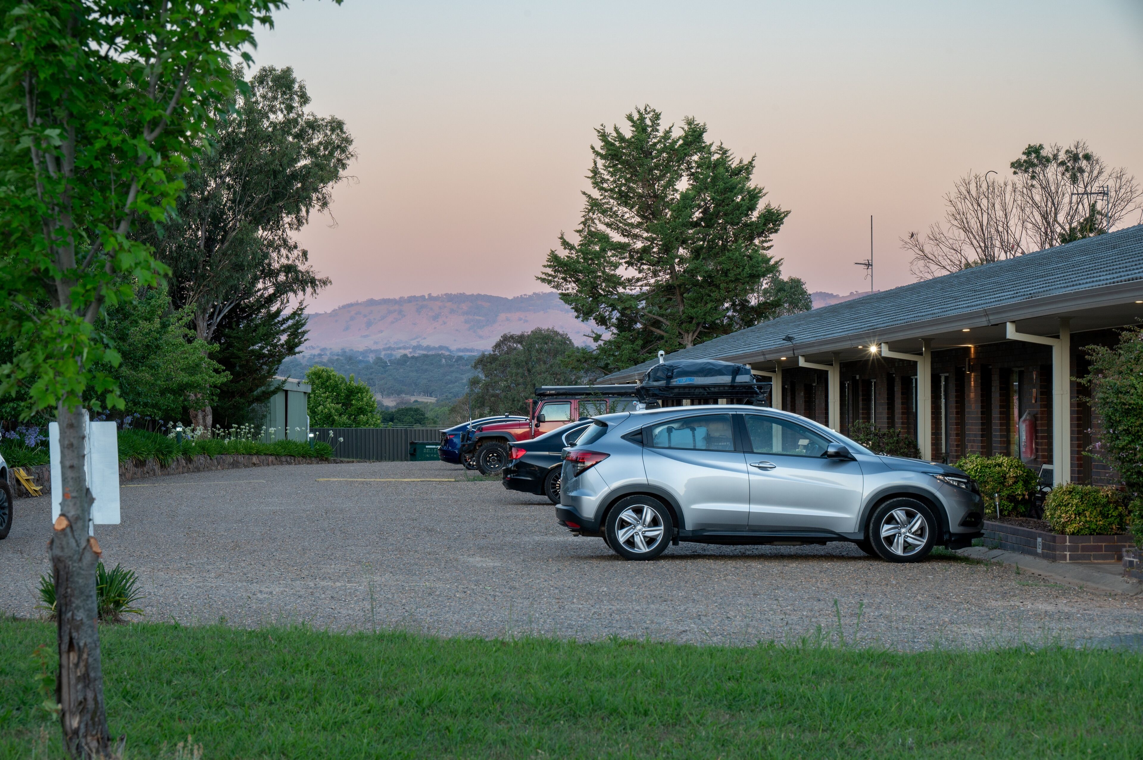 Un estacionamiento con varios autos, un edificio de techo inclinado y montañas al fondo.