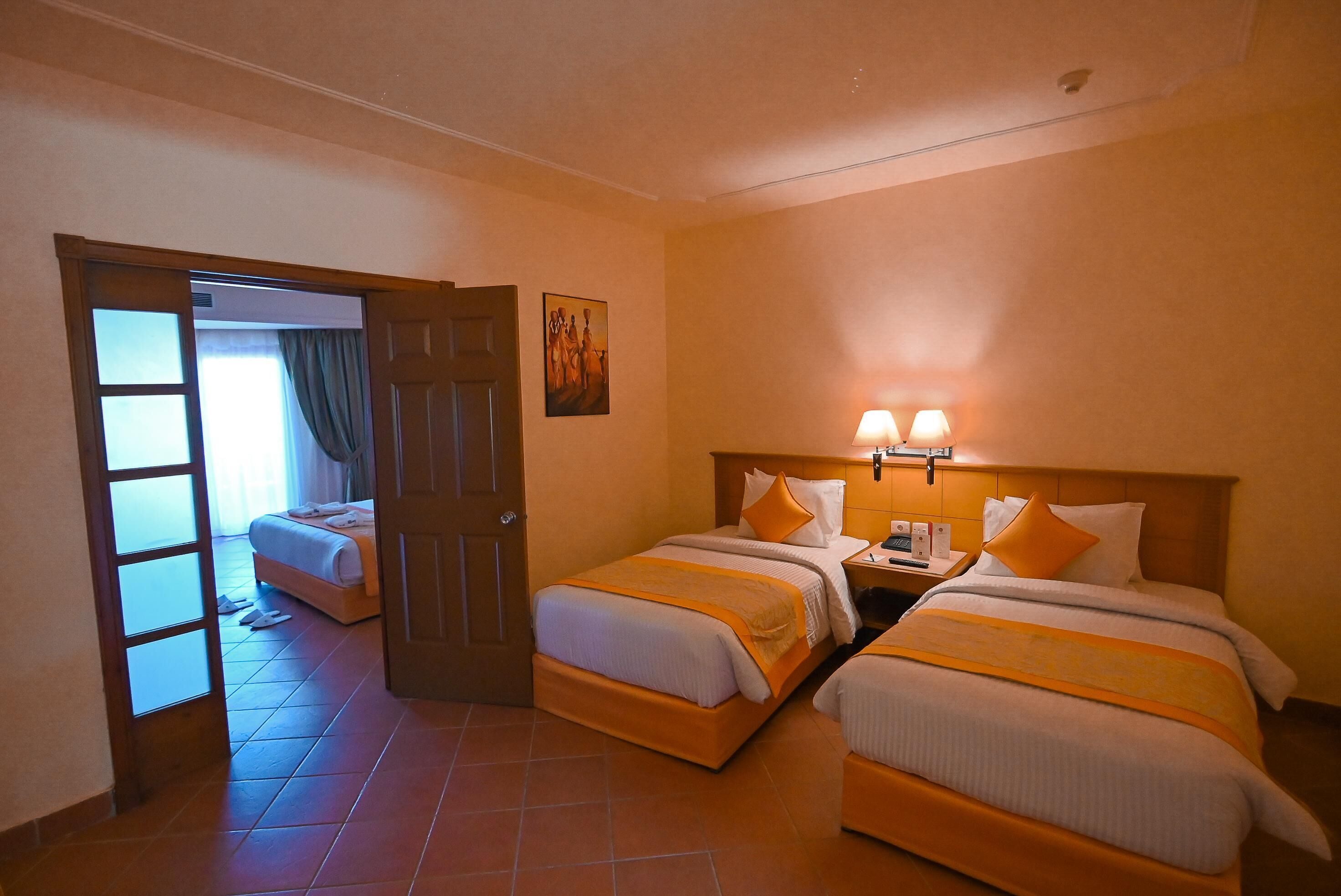 Une chambre d’hôtel avec deux lits, une porte donnant sur une autre pièce, une table de chevet avec une lampe et un tableau au mur.