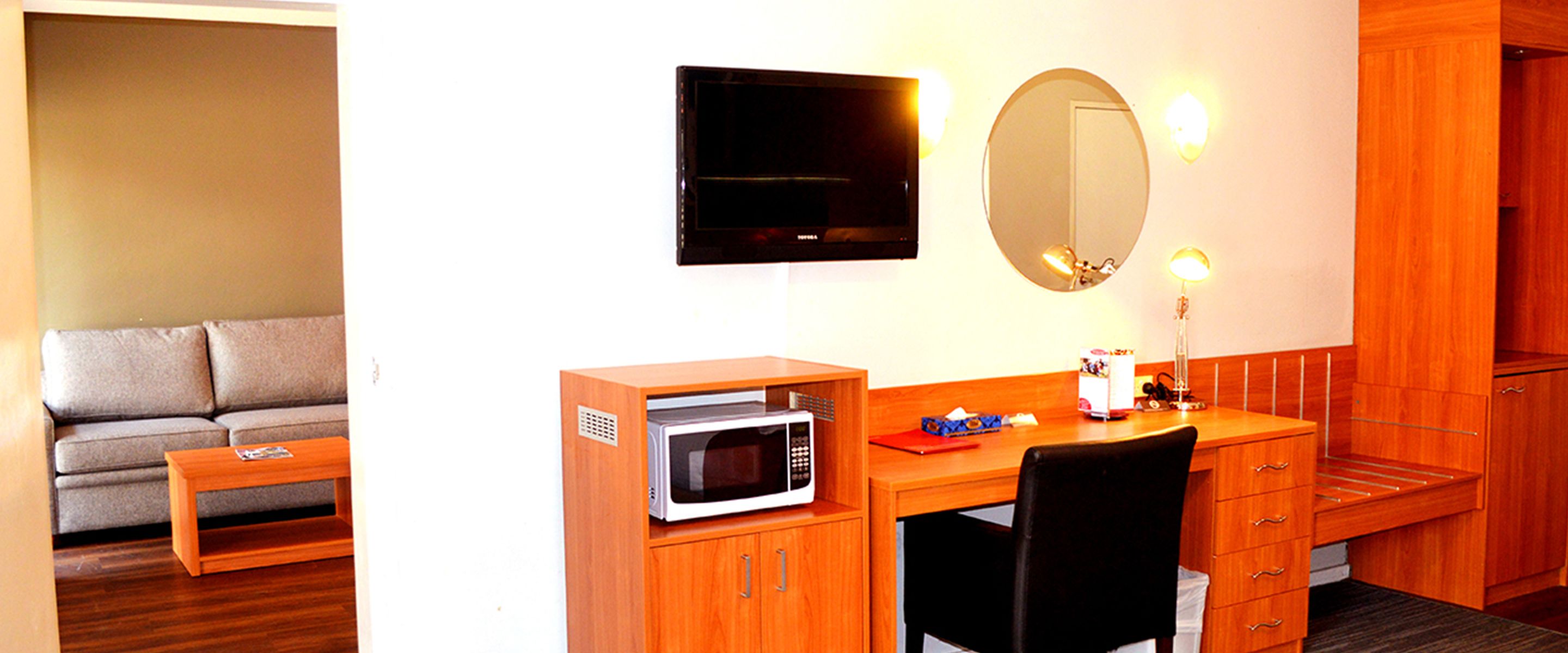 Una habitación compacta con escritorio de madera, microondas, pequeña silla, televisor y espejo.
