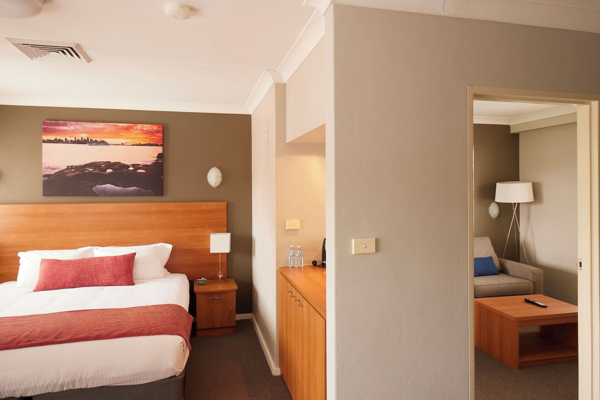 Habitación de hotel con cama, mesitas de noche, lámpara, un cuadro y vista a otra habitación.