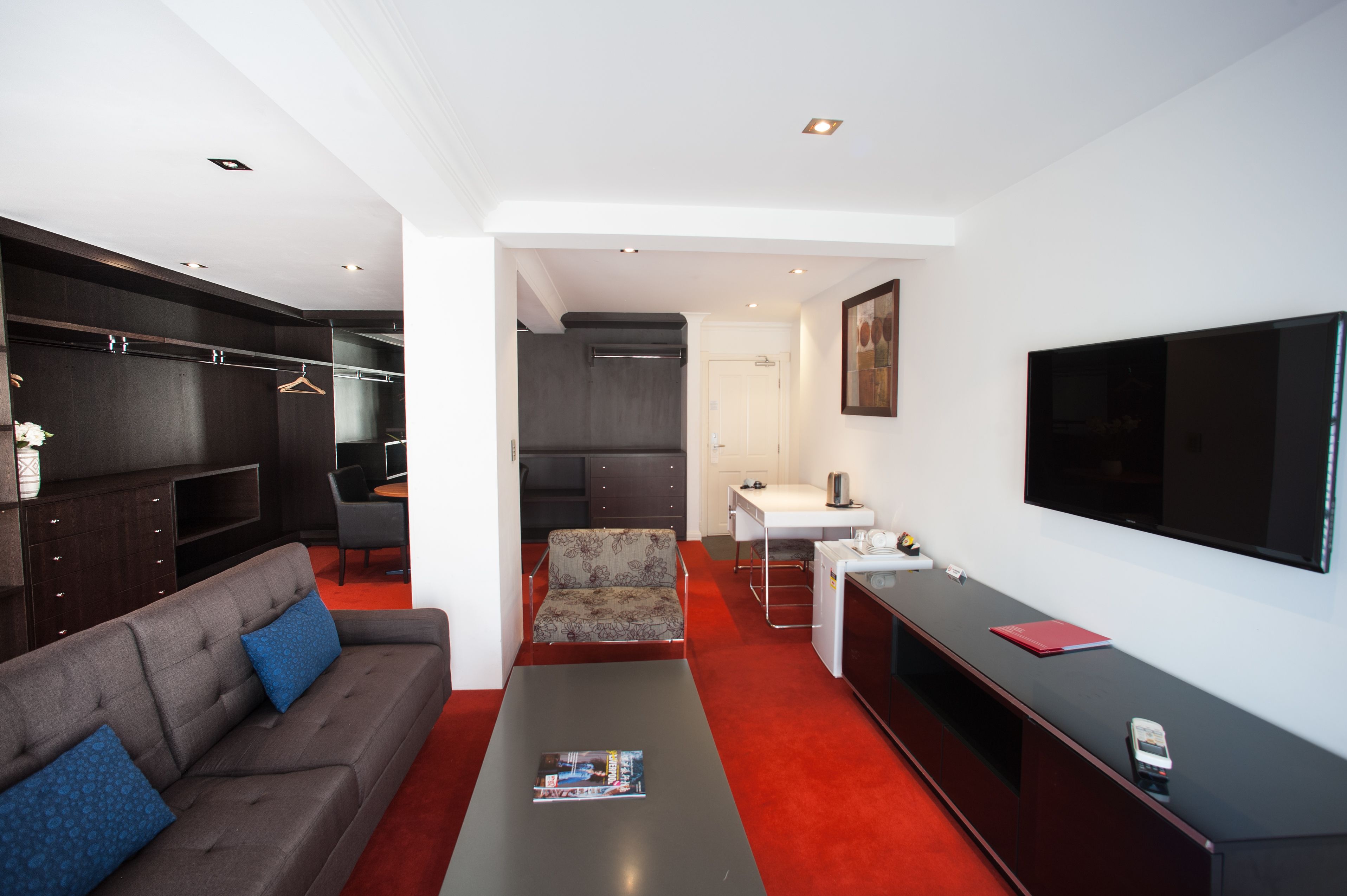 Une chambre d’hôtel moderne avec un canapé gris, une petite table, un téléviseur, un bureau et une armoire.