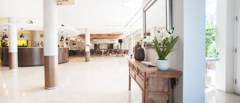 Um hall amplo com balcão de recepção, área de estar e sinalização indicando serviços como restaurante, sala de conferências, bar, jogos e um caixa eletrônico.