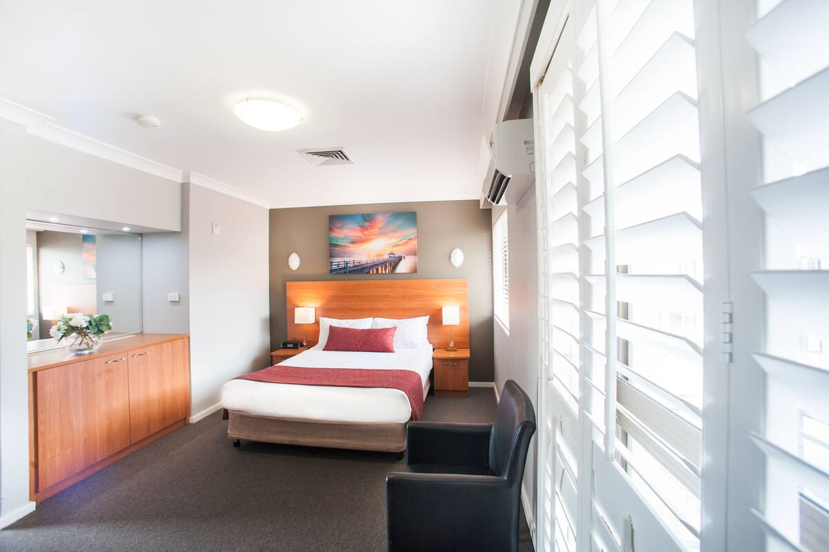 A hotel room with a bed, bedside tables, a desk, a chair, a mirror, and a painting on the wall.