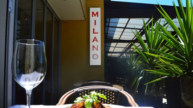 A dining table with a plate of food and a wine glass, a wicker chair, and a sign that reads "MILANO". - Ramada Hotel & Suites Sydney Cabramatta (Cabramatta)