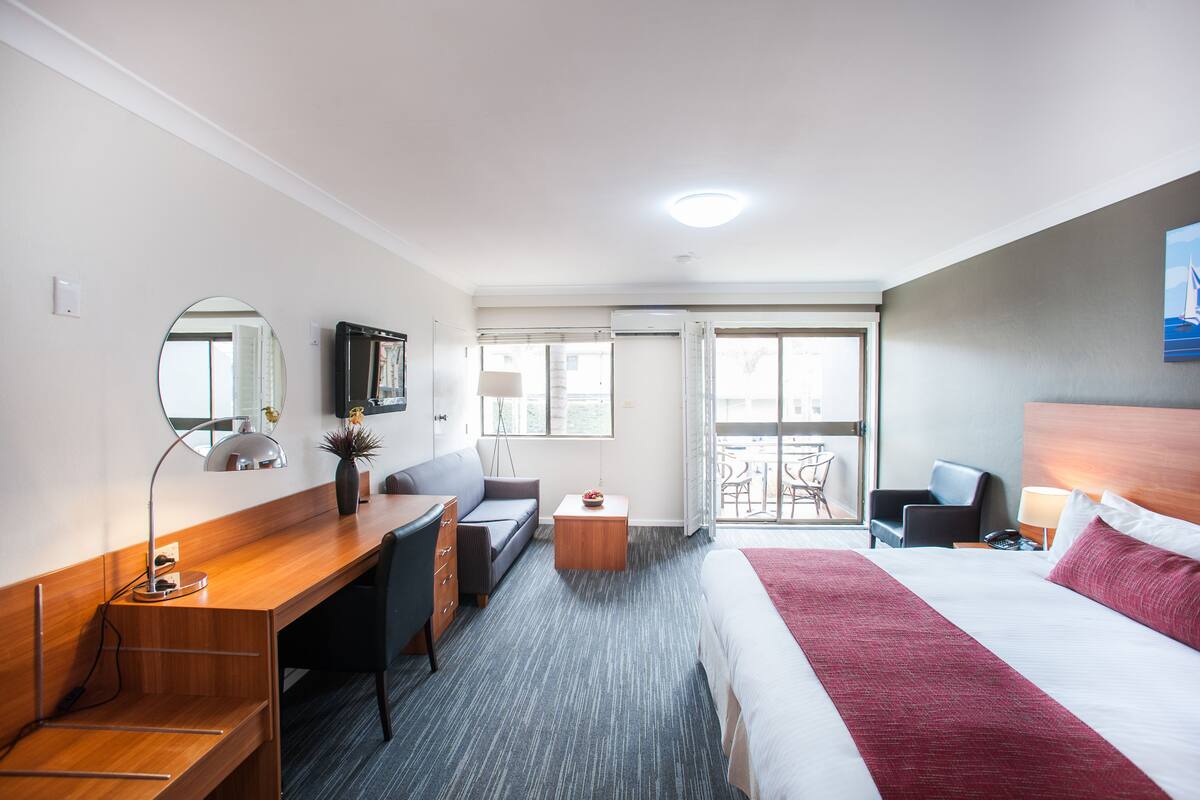 A hotel room with a bed, desk, chair, and a small table with a lamp.