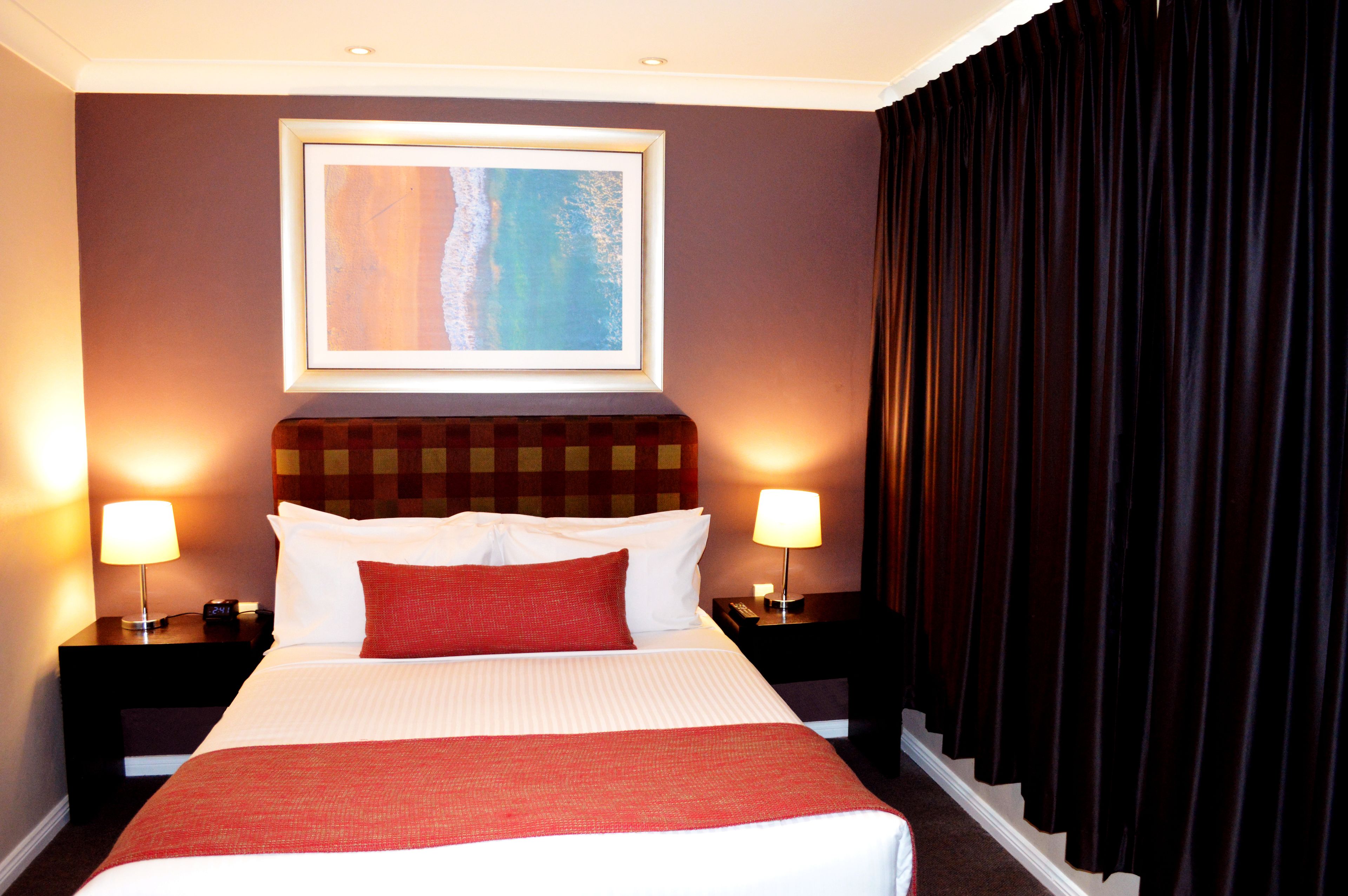 A hotel room with a large bed, two bedside lamps, a framed artwork above the bed, and dark curtains on the window.
