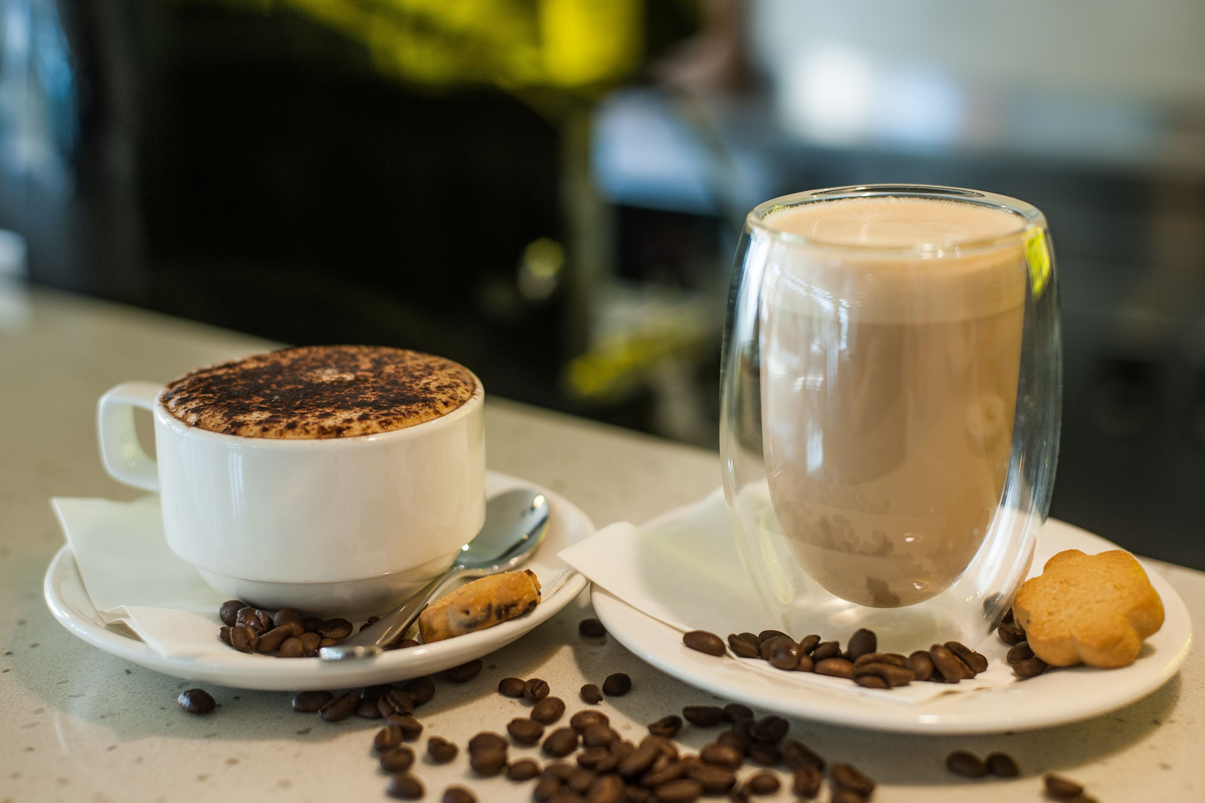 Two cups of coffee, one with a frothy top, and a glass of iced coffee on a table with scattered coffee beans and a biscuit.