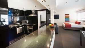 A modern kitchen with stainless steel appliances, a dining area with a sofa and a TV, and a living room with a red pillow. - Ramada Hotel & Suites Sydney Cabramatta (Cabramatta)