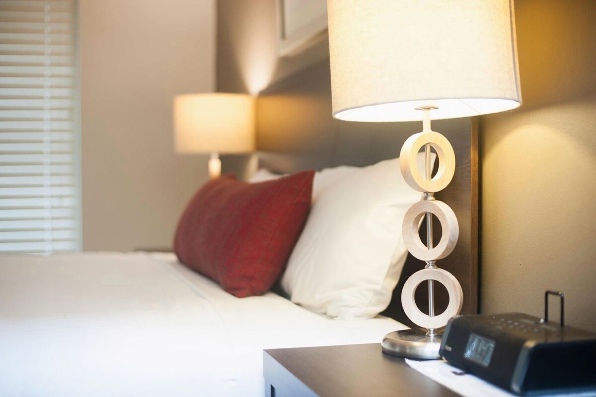 A hotel room with a bed, a nightstand with a lamp, and a digital clock.