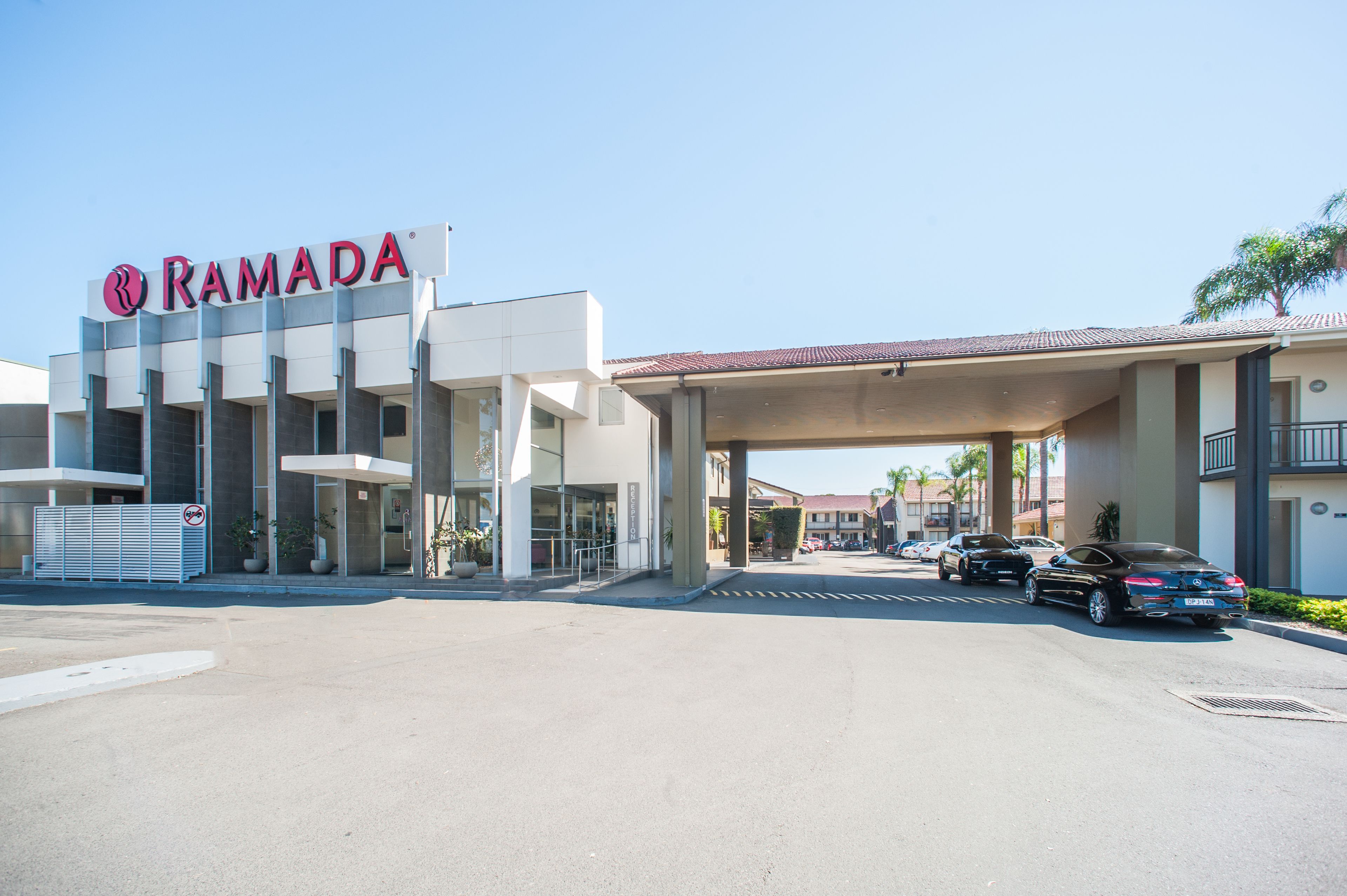 Ein modernes Gebäude mit einem 'RAMADA'-Schild, einem überdachten Parkbereich und blauem Himmel.