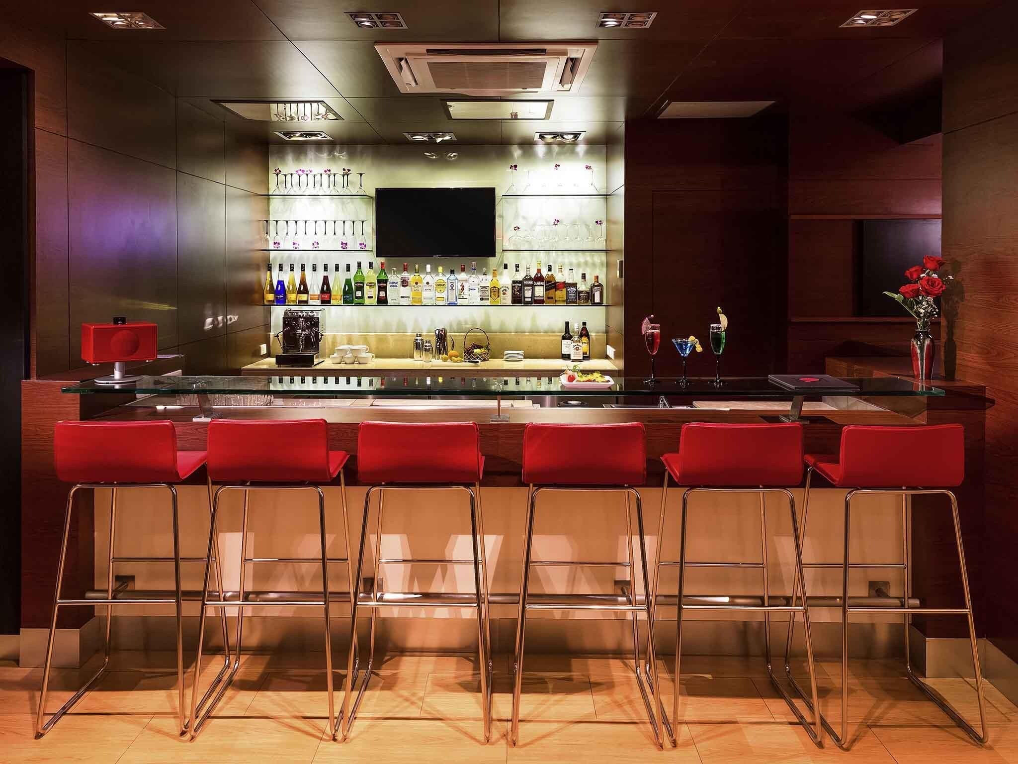 Zona de bar con mostrador de vidrio, taburetes rojos y una variedad de botellas y vasos en estantes.