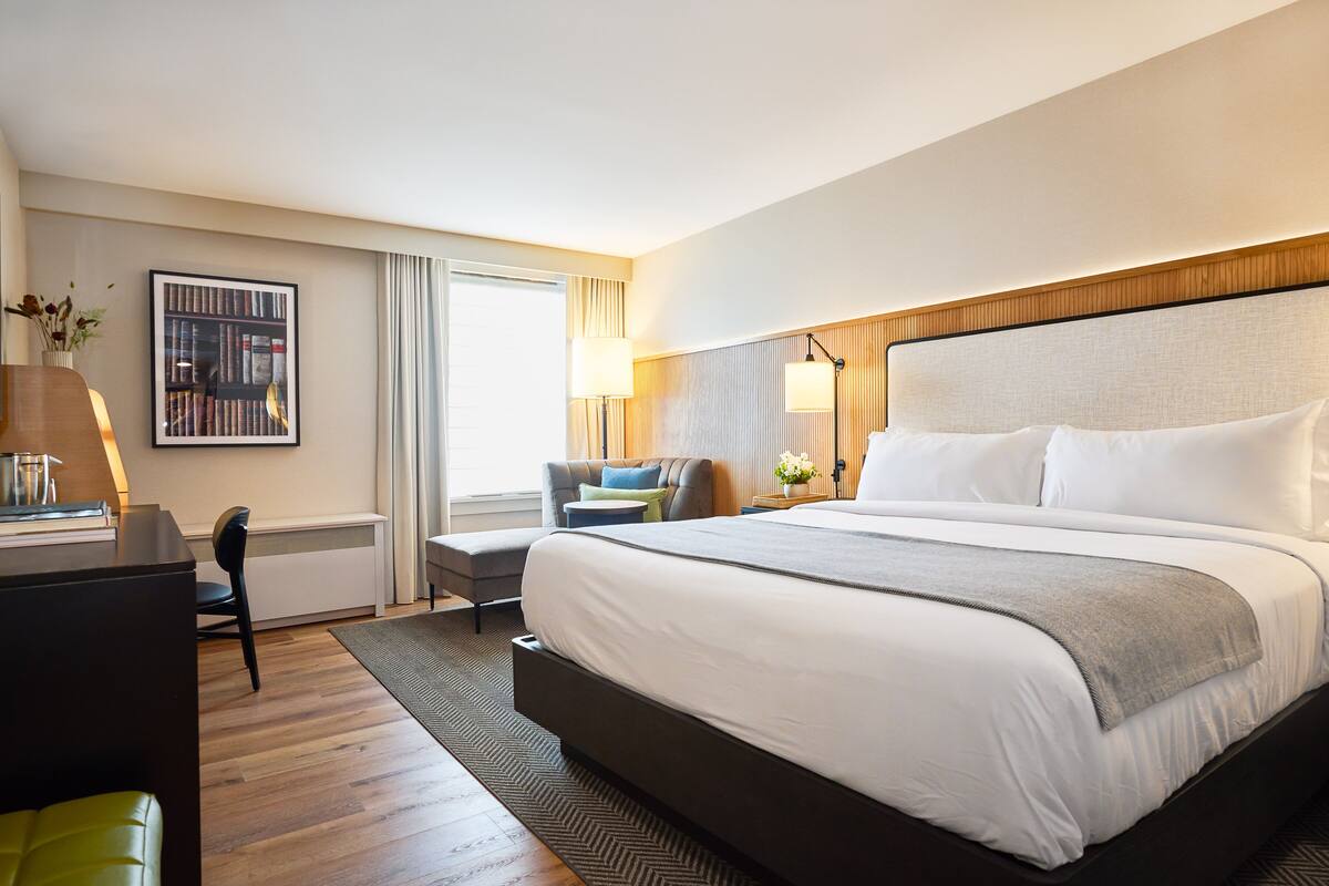 A hotel room with a large bed, a desk with a lamp, a chair, a bookshelf, and a window with curtains.