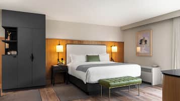 A modern hotel room with a large bed, bedside lamps, a green bench, a wardrobe, and a desk with a lamp.