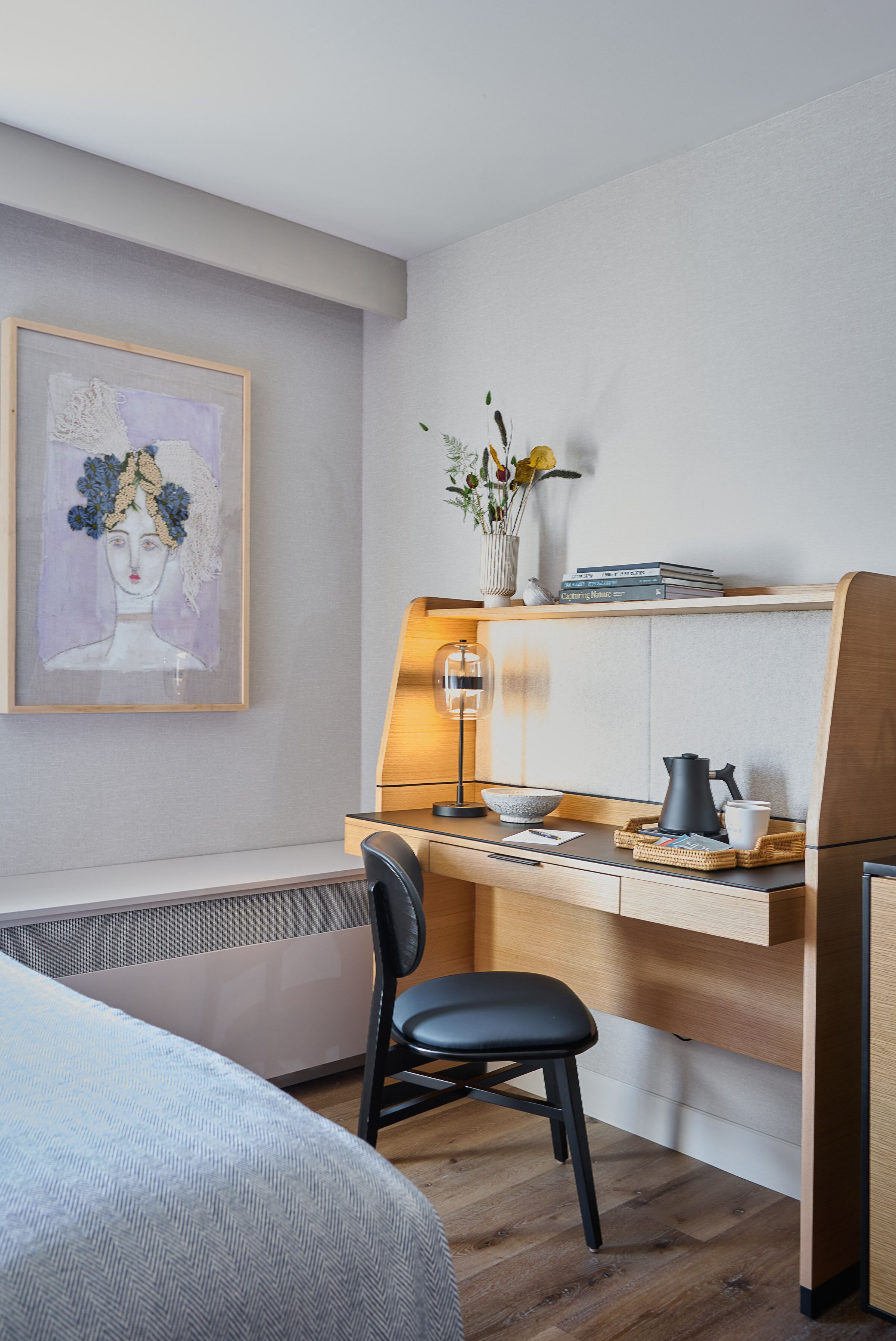 Une chambre moderne avec un bureau en bois, une chaise, un lit et une étagère murale ornée de livres et d’un vase de fleurs.