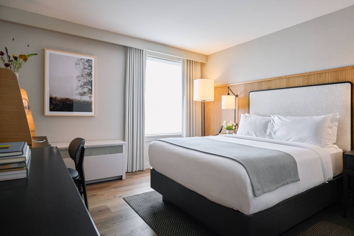 A hotel room with a large bed, a desk with a chair, a framed picture on the wall, and a window with curtains.
