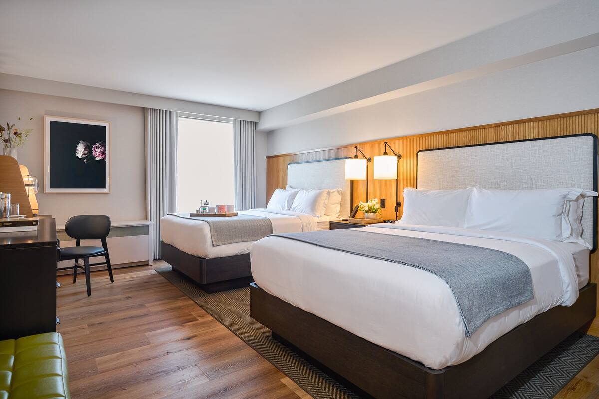 A hotel room with two beds, a desk, and a chair. There is a framed picture on the wall and a window with curtains.