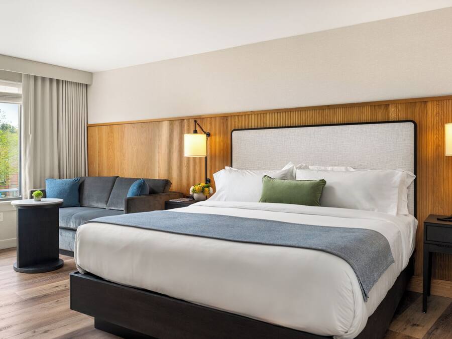 A modern hotel room with a large bed, a sofa, a side table, and a lamp.
