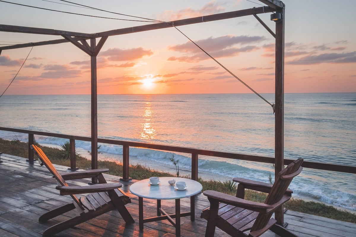 Terraza de madera con dos mecedoras y una mesita pequeña, con vista al océano al atardecer.