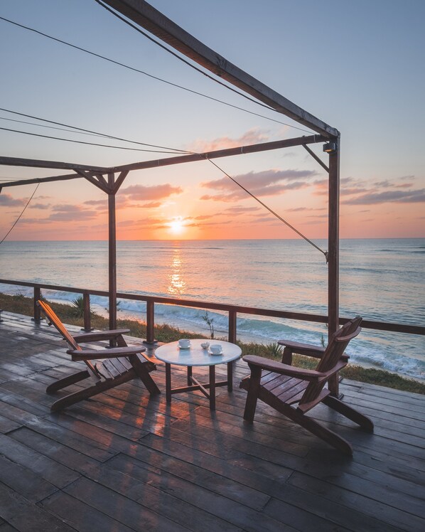 Terraza de madera con dos mecedoras y una mesita pequeña, con vista al océano al atardecer.