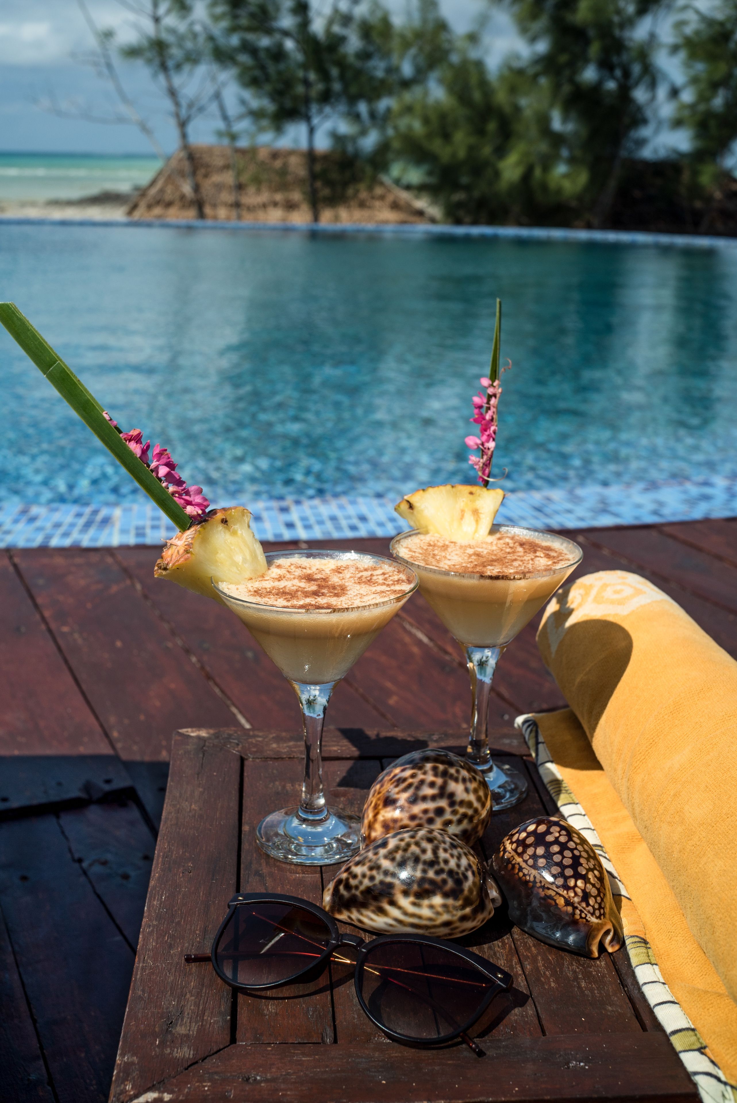 Una terraza de madera con dos cócteles tropicales adornados con piña y una flor, un par de gafas de sol y dos conchas marinas.