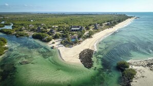 An aerial view of a coastal resort with multiple thatched-roof cottages, a sandy beach, and clear turquoise waters.