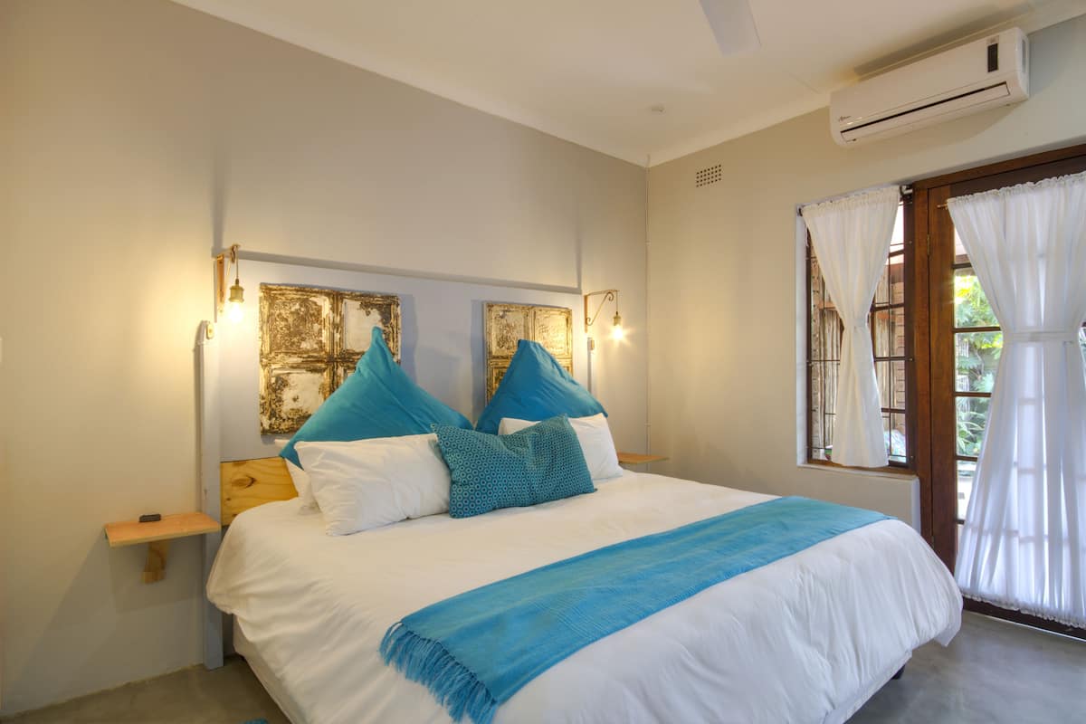 Quarto com cama, mesa de cabeceira, ar condicionado fixado na parede e janela com cortinas.