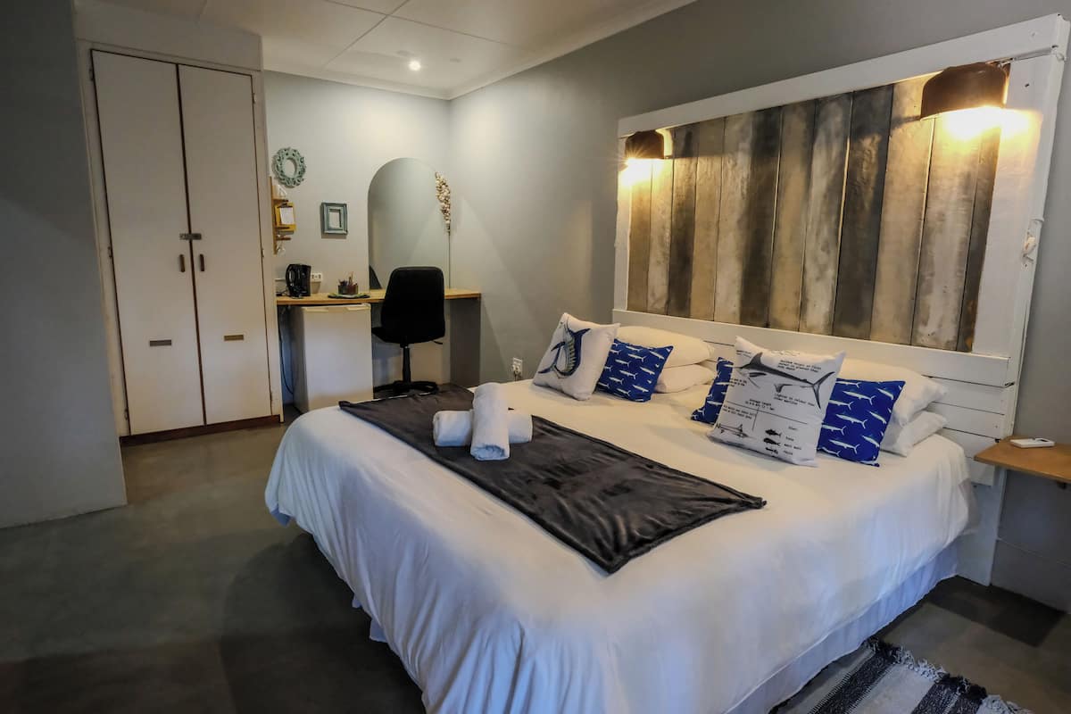 Cama bem arrumada com roupa de cama branca e almofadas estampadas em azul, manta preta e cabeceira de madeira com luminária.
