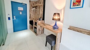 Ein Hotelzimmer mit blauer Tür, einem Holztisch mit Lampe, einem kleinen Hocker und einem an der Wand befestigten Kleiderhaken.