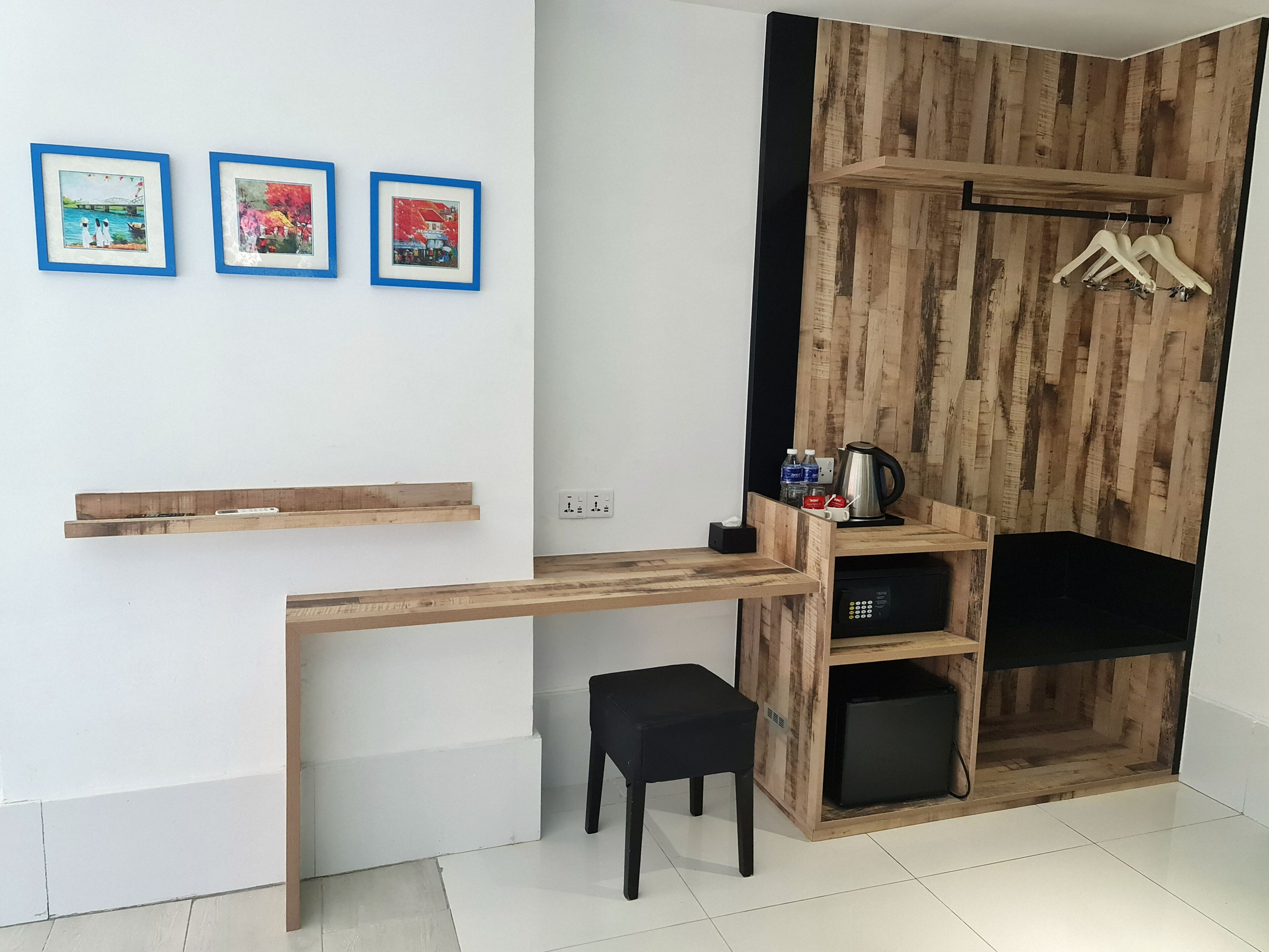 Una habitación compacta con un escritorio de madera, una silla negra y un taburete pequeño. Hay tres cuadros enmarcados en la pared, una estantería de madera con una tetera y botellas, y un perchero de madera con perchas.