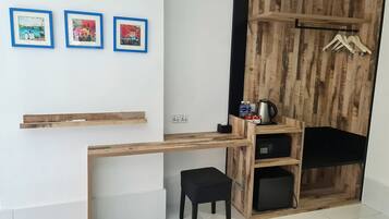 Una habitación compacta con un escritorio de madera, una silla negra y un taburete pequeño. Hay tres cuadros enmarcados en la pared, una estantería de madera con una tetera y botellas, y un perchero de madera con perchas.