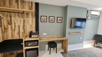 Una habitación moderna con un escritorio de madera, una silla de oficina negra, un televisor instalado en la pared y un refrigerador pequeño.