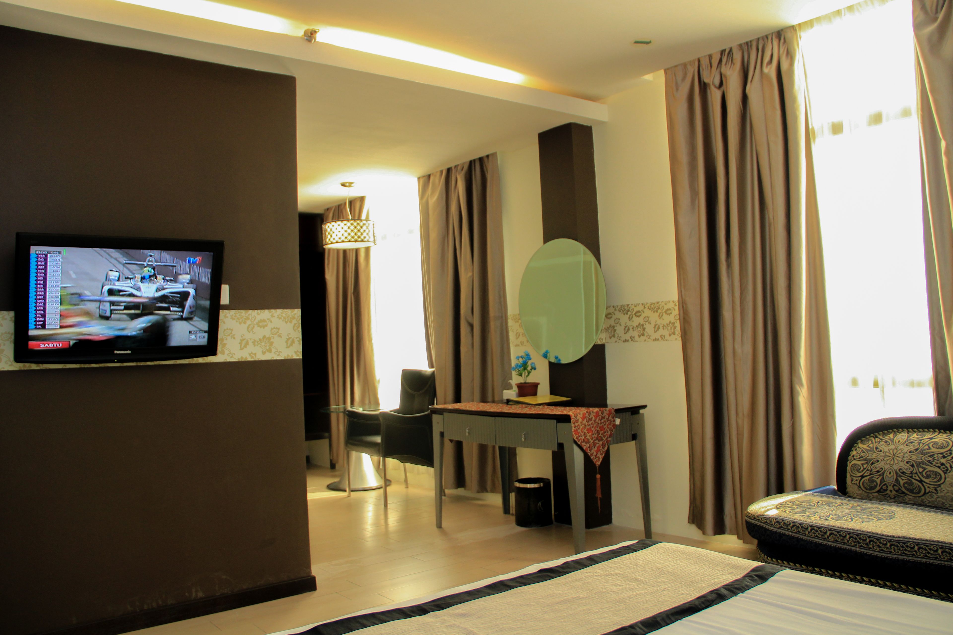 A hotel room with a television displaying a racing game, a dining table with chairs, a sofa, and a large mirror.