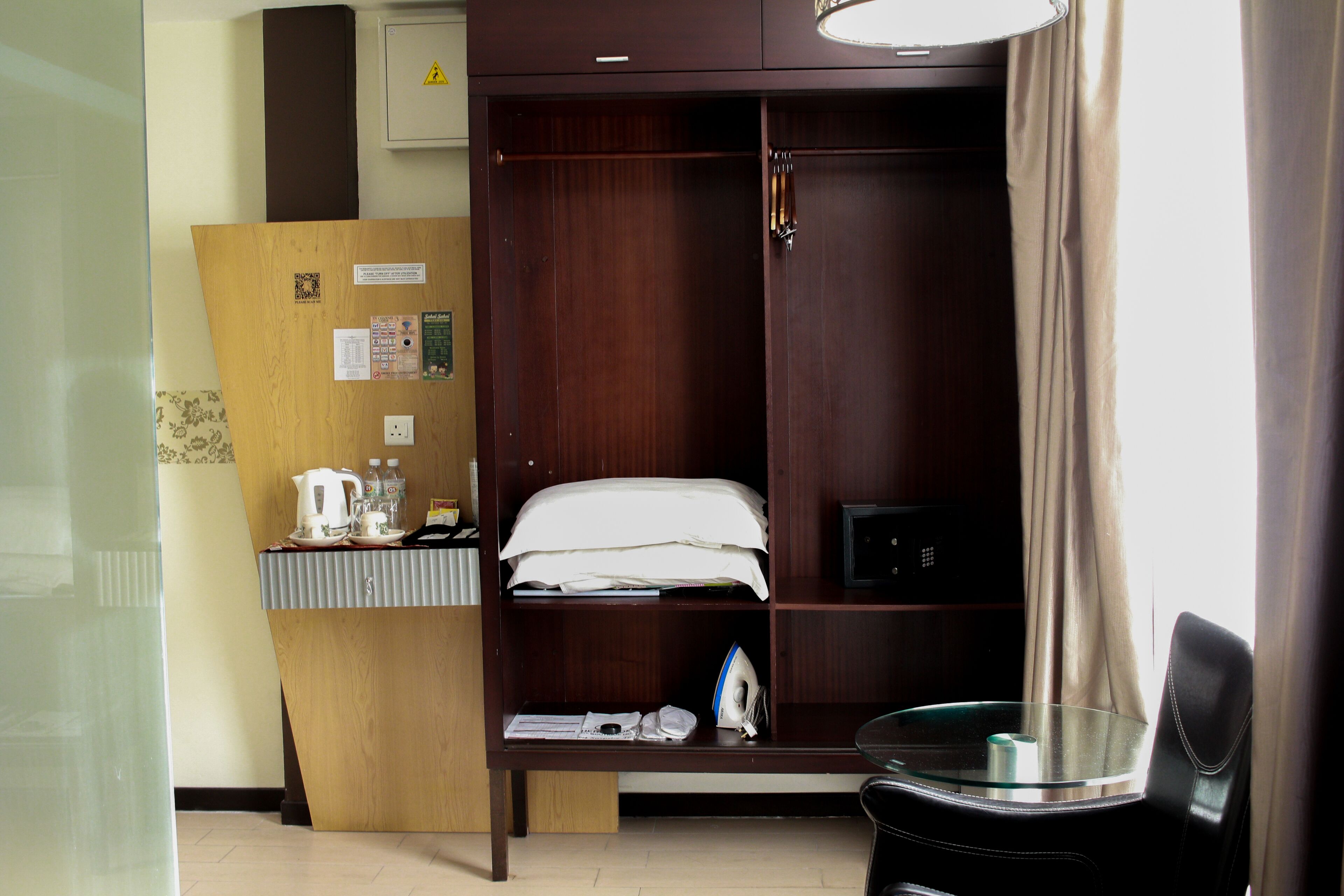 A hotel room with a wooden wardrobe, a small desk with a kettle and cups, a bed with a white comforter, and a glass table with a chair.