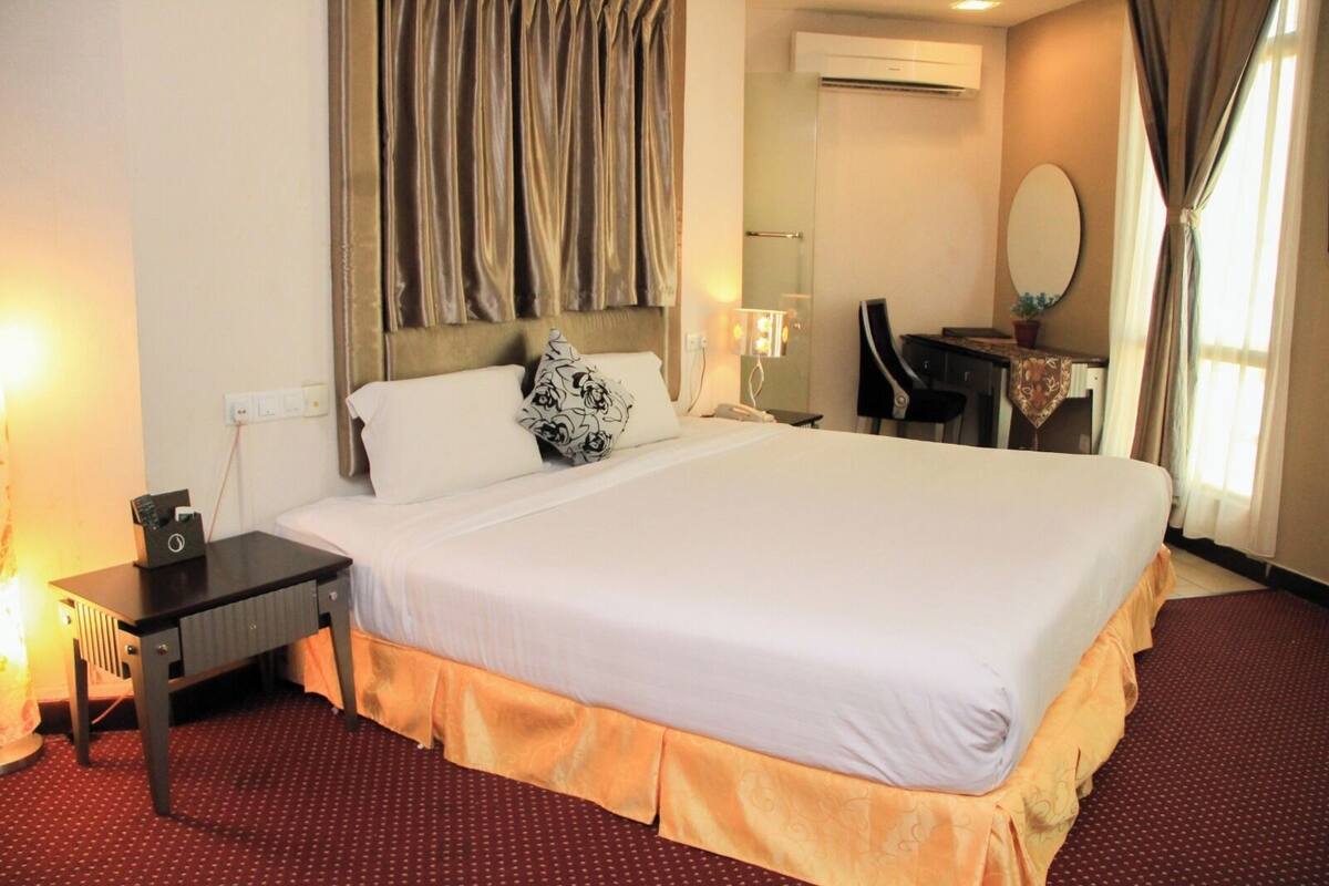 Una habitación de hotel con una cama grande, una mesita de noche con lámpara, un escritorio con silla, un espejo y una ventana con cortinas.