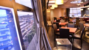 Un moderno comedor con mesas y sillas, un ventanal con cortinas y vistas a un paisaje urbano.