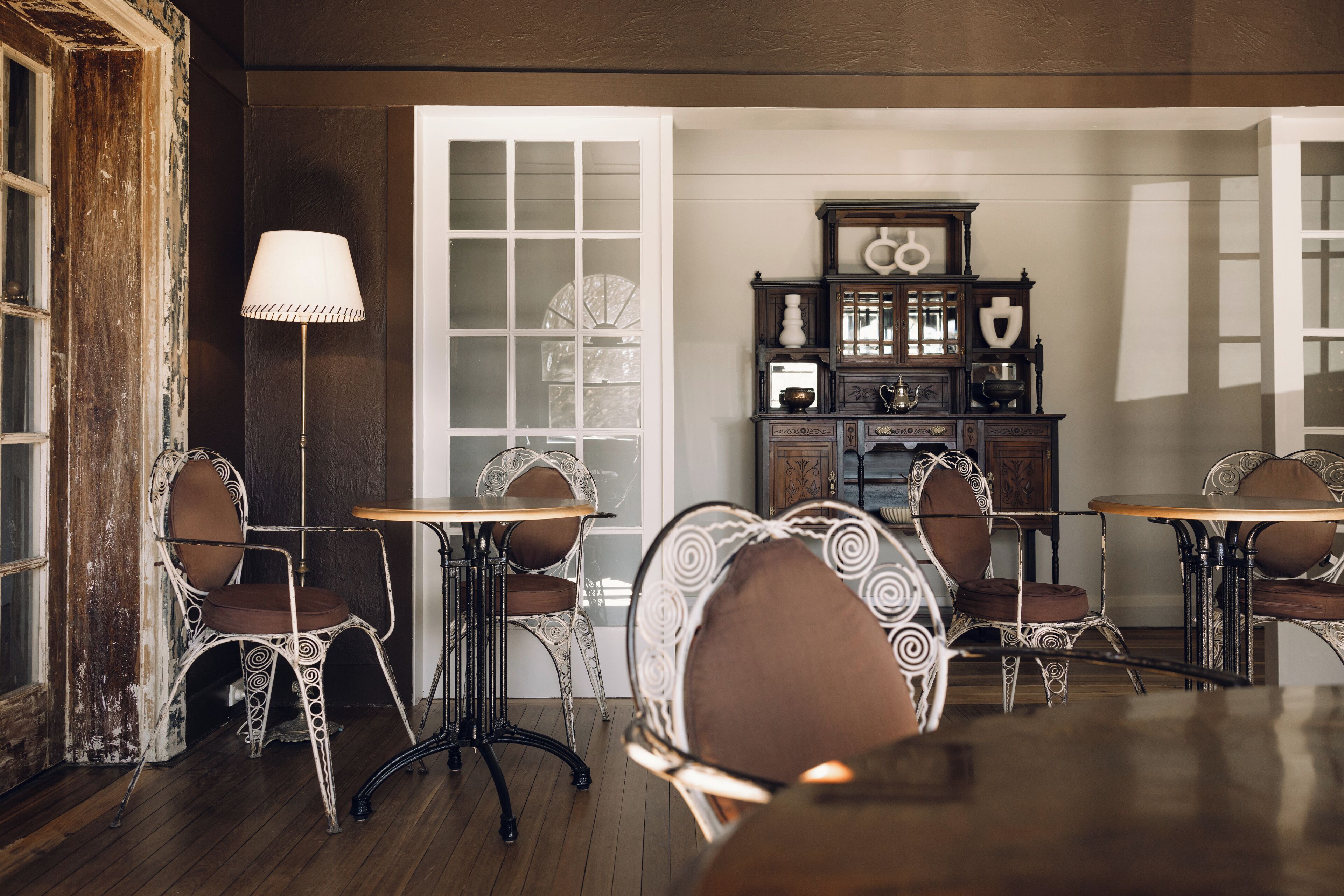 A dining area with ornate chairs and tables, a vintage lamp, and a wooden cabinet with glass doors.