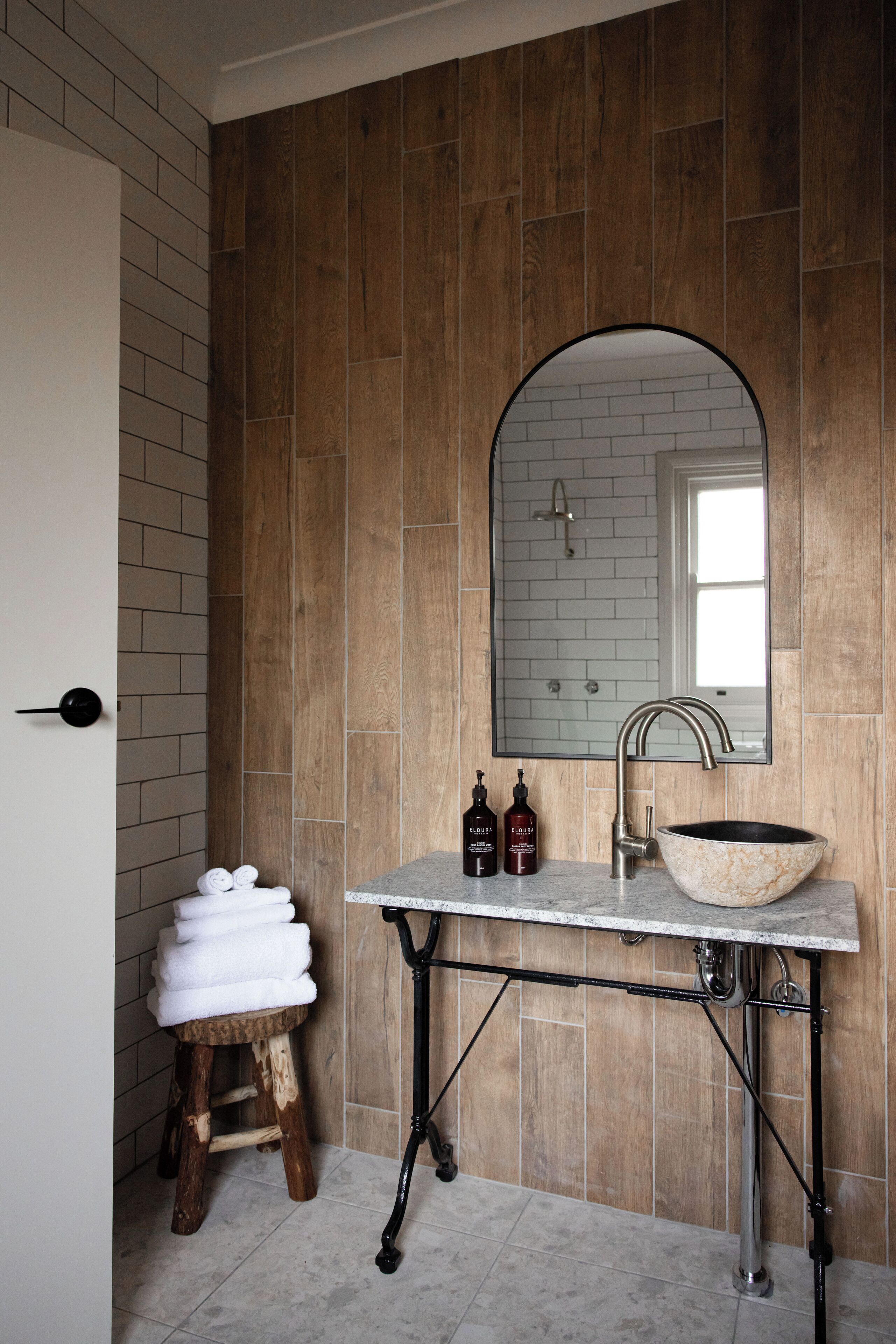 A bathroom with a wooden wall, a white sink with a vessel basin, a mirror, and a wooden stool with towels.