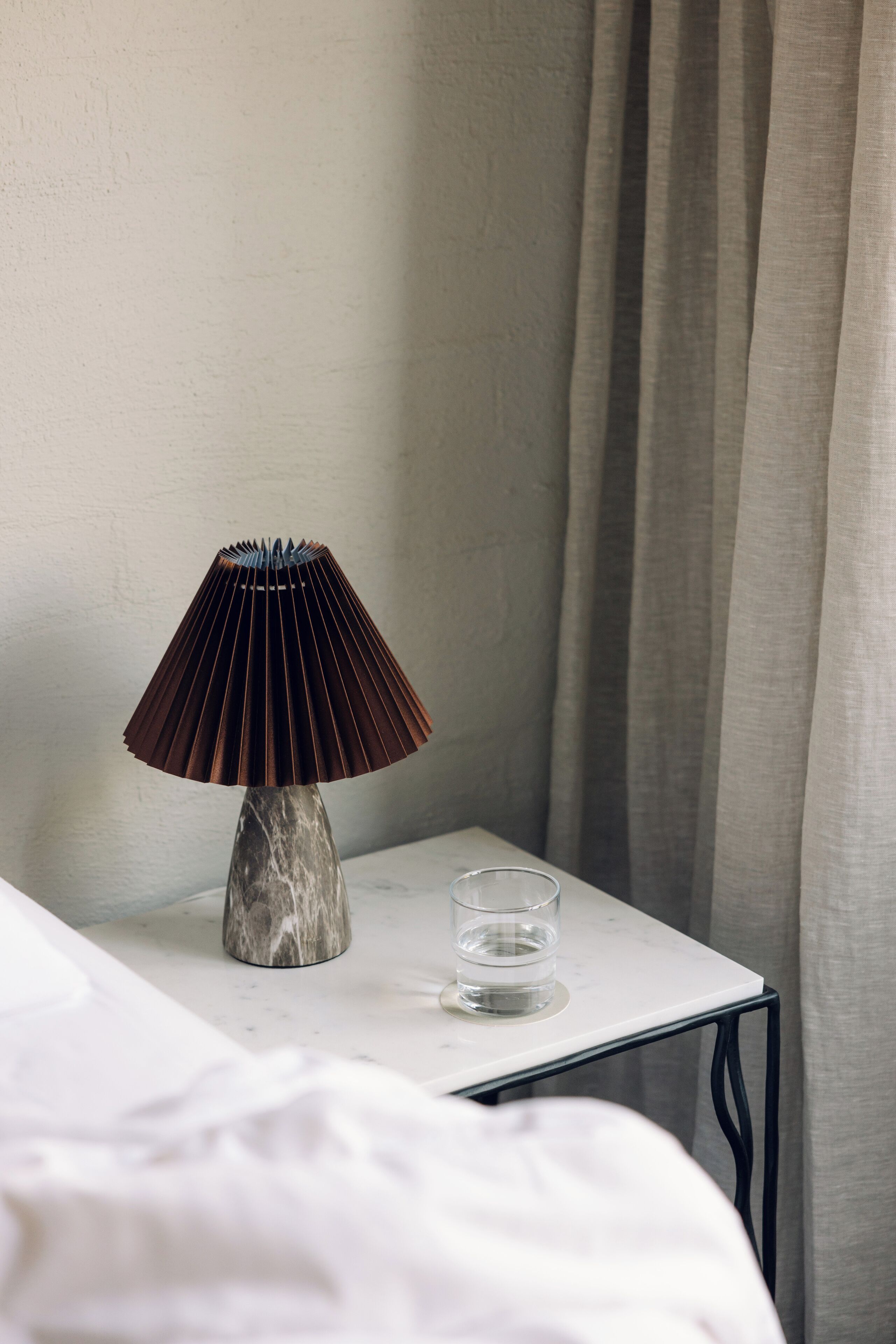 A bed with white linens, a marble nightstand with a lamp, and a glass of water.