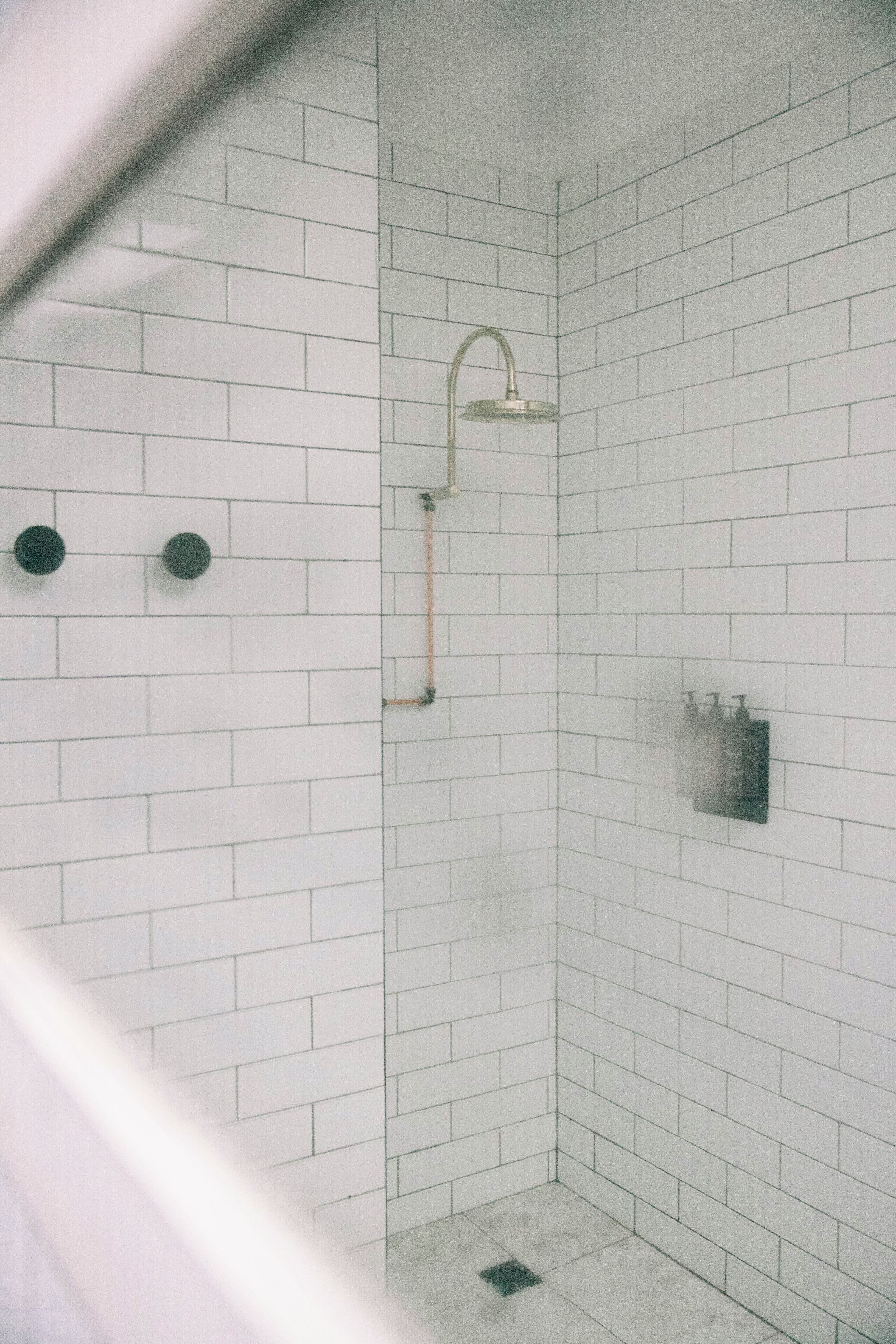 Une douche carrelée de carreaux blancs, dotée d’un pommeau de douche à main, d’un porte-serviettes et d’un distributeur de savon.