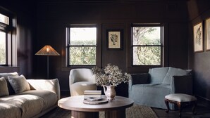 A living room with a sofa, armchair, and ottoman, a coffee table with a vase and books, and large windows with a view of trees. - Links House (Burradoo)
