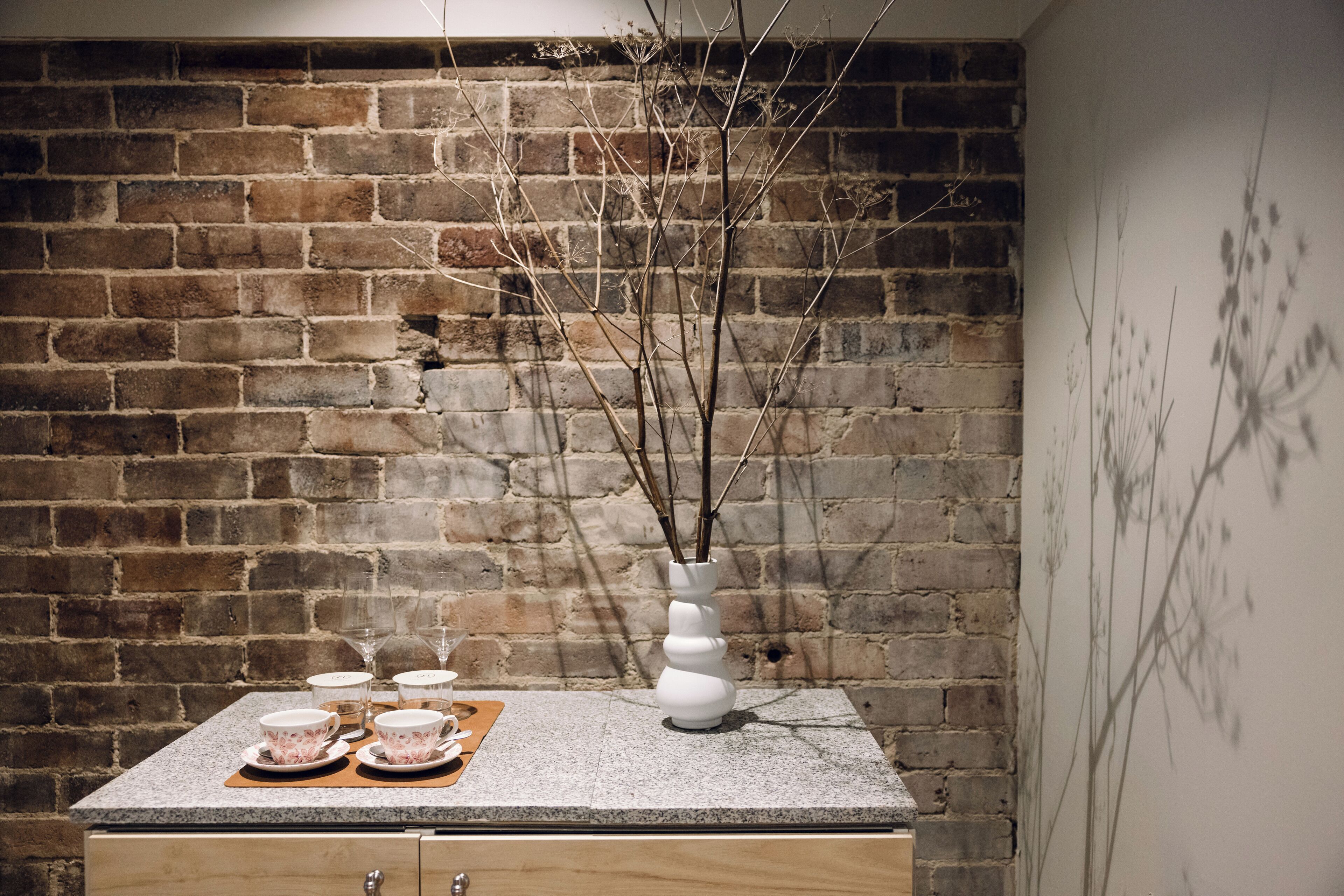 A dining area with a brick wall, a table set with white plates, cups, and a vase with branches.