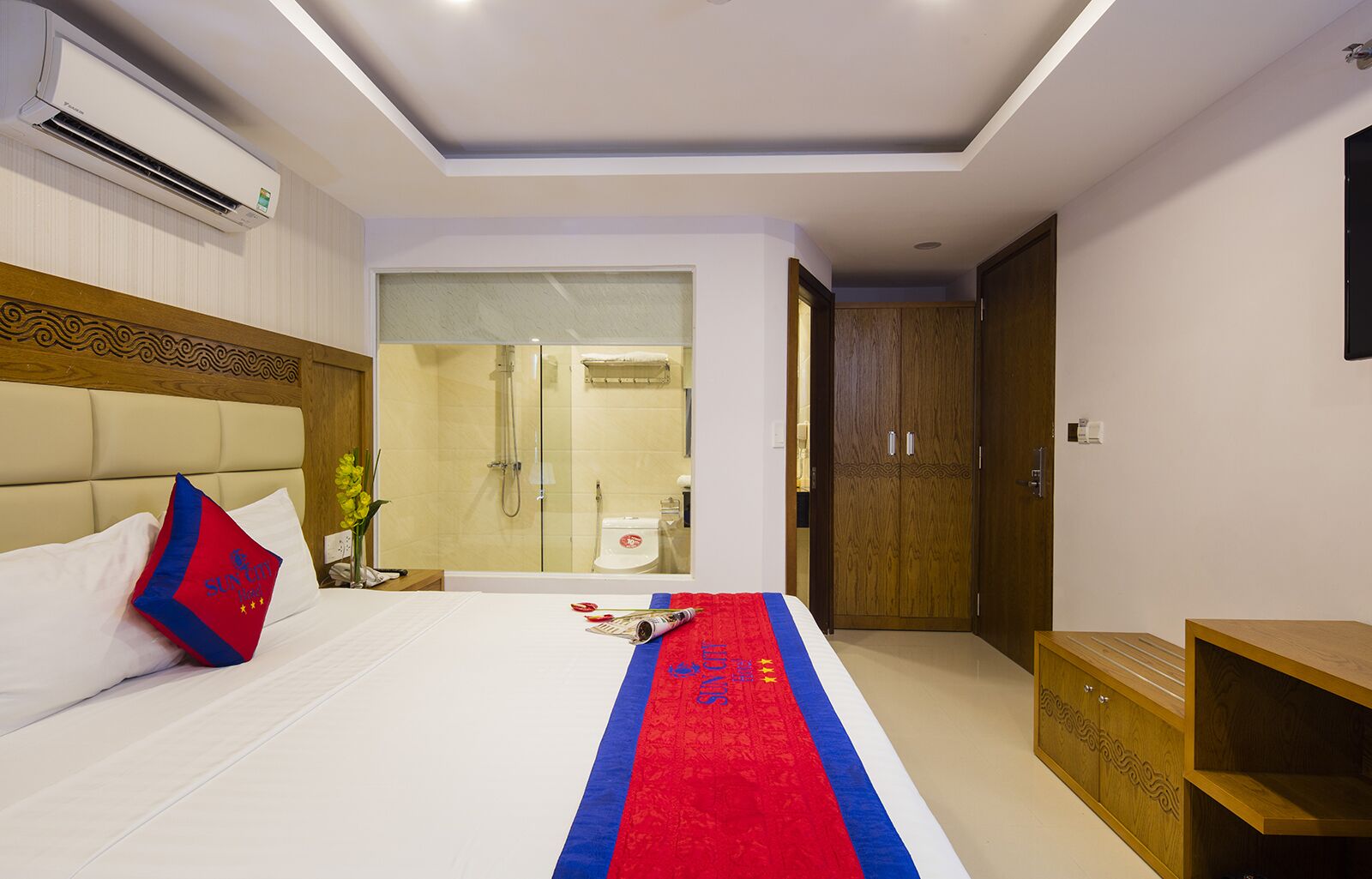 A hotel room with a bed, a wooden headboard, a red and blue decorative runner, a wooden nightstand, a wardrobe, and a shower area visible through a glass door.