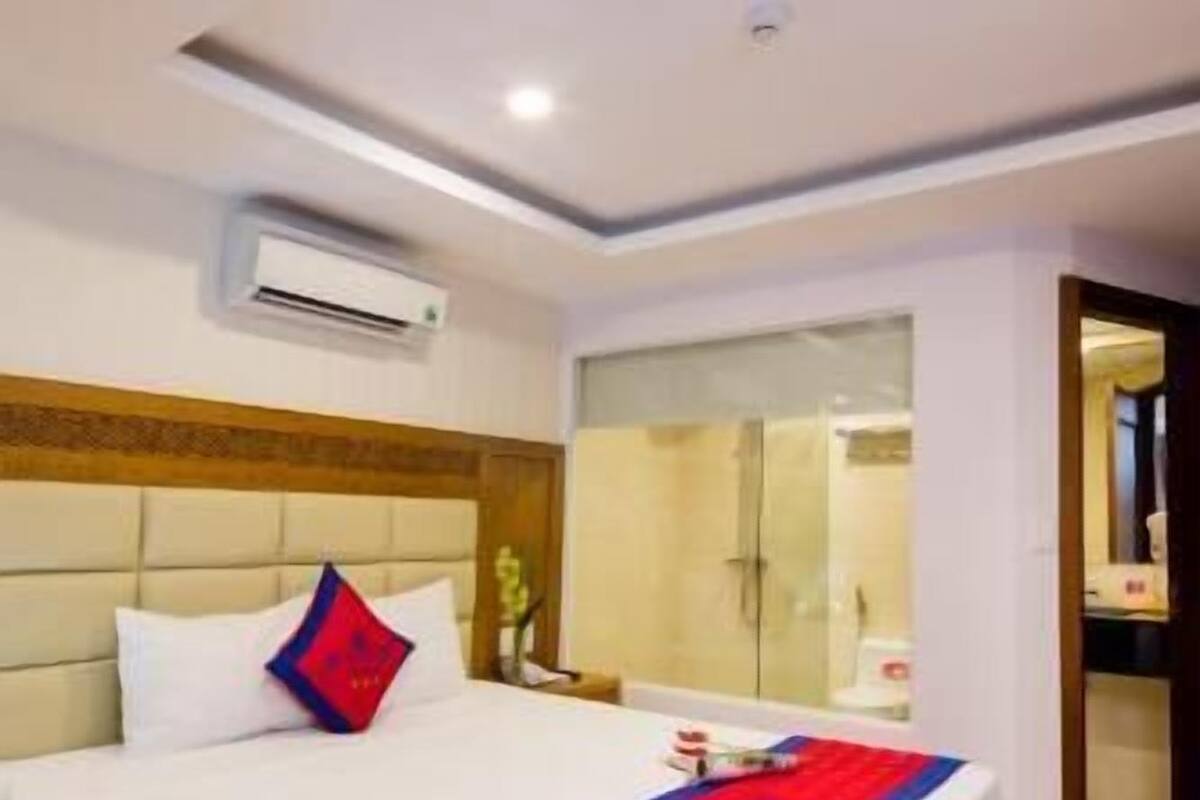 A hotel room with a large bed, a wooden headboard, an air conditioning unit, a bathroom with a glass door, and a visible hallway.