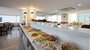 Un bufé con diversas opciones de comida, una zona de estar con sillas y un amplio ventanal que proporciona luz natural.