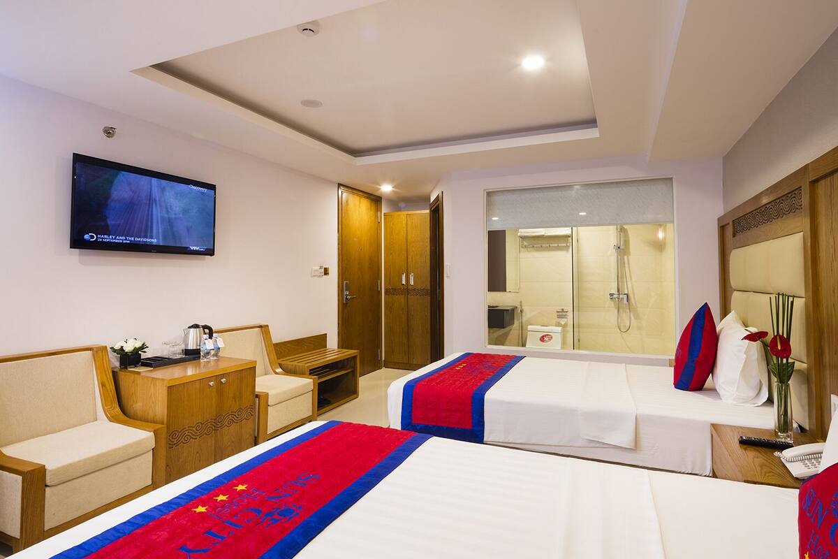 A hotel room with two beds, a TV, a desk, a chair, a wardrobe, and a bathroom visible through a glass door.