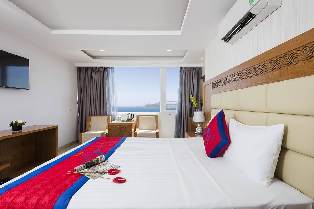 A hotel room with a large bed, a desk, a chair, a TV, and a view of the sea.