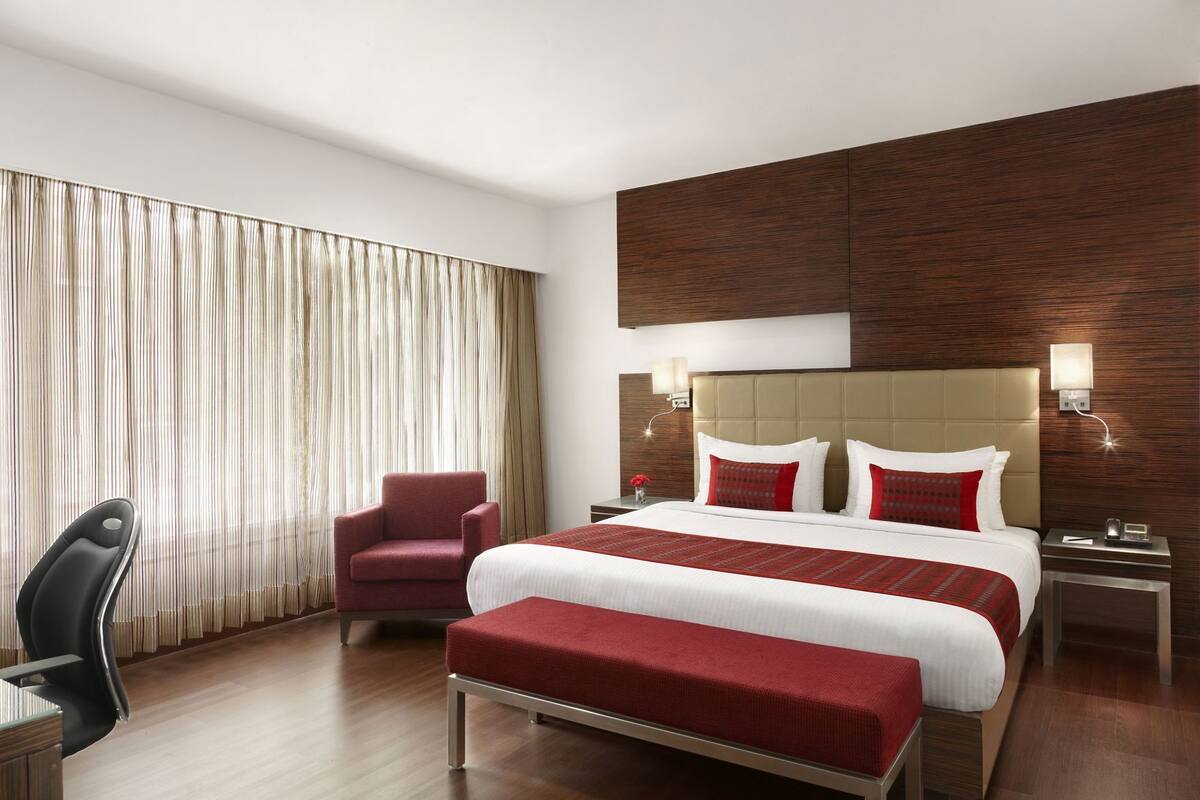 A hotel room with a large bed, a red armchair, a desk, and a window with curtains.