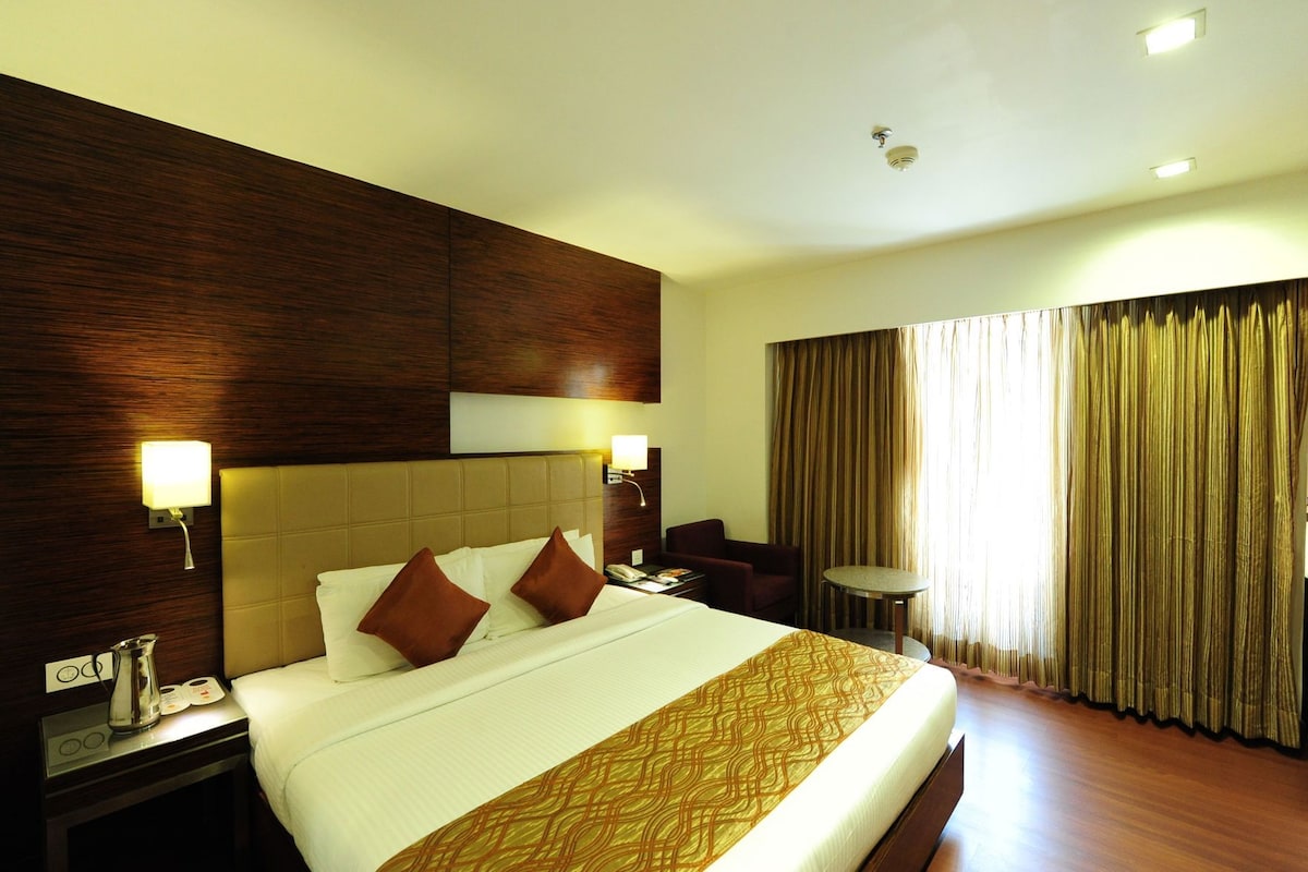 Ein Hotelzimmer mit einem großen Bett, einem Nachttisch, einem Schreibtisch, einem Sessel, einem kleinen Tisch und einem Fenster mit Vorhängen.